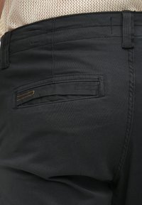 Μαύρο παντελόνι από λείο ύφασμα με εμφανείς ραφές και μια μοναδική πίσω τσέπη με πορτοκαλί λεπτομέρεια.