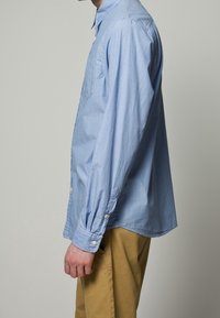 Vaaleansininen raidallinen nappipaita, jossa on pitkät hihat, kaulus ja yksittäinen rintatasku, yhdistettynä vaaleanruskeisiin housuihin.