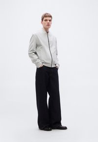 Ανοιχτό γκρι μπουφάν μπομπέρ με ριμπ μανσέτες, μαύρο φερμουάρ και πλαϊνές τσέπες, σε συνδυασμό με φαρδιά σκουρόχρωμα τζιν και μαύρα παπούτσια.