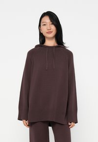 Jovem mulher com cabelo preto longo, vestindo um hoodie de malha solto e escuro, com calças a condizer, em pé contra um fundo branco liso.