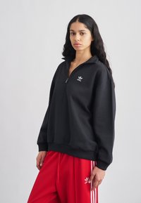 Juodas pusiau užtraukiamas vilnonis džemperis su išsiuvinėtu baltu logotipu, derinamas su raudonomis sportinėmis kelnėmis su baltomis šoninėmis juostelėmis ir atitinkančiu logotipu.