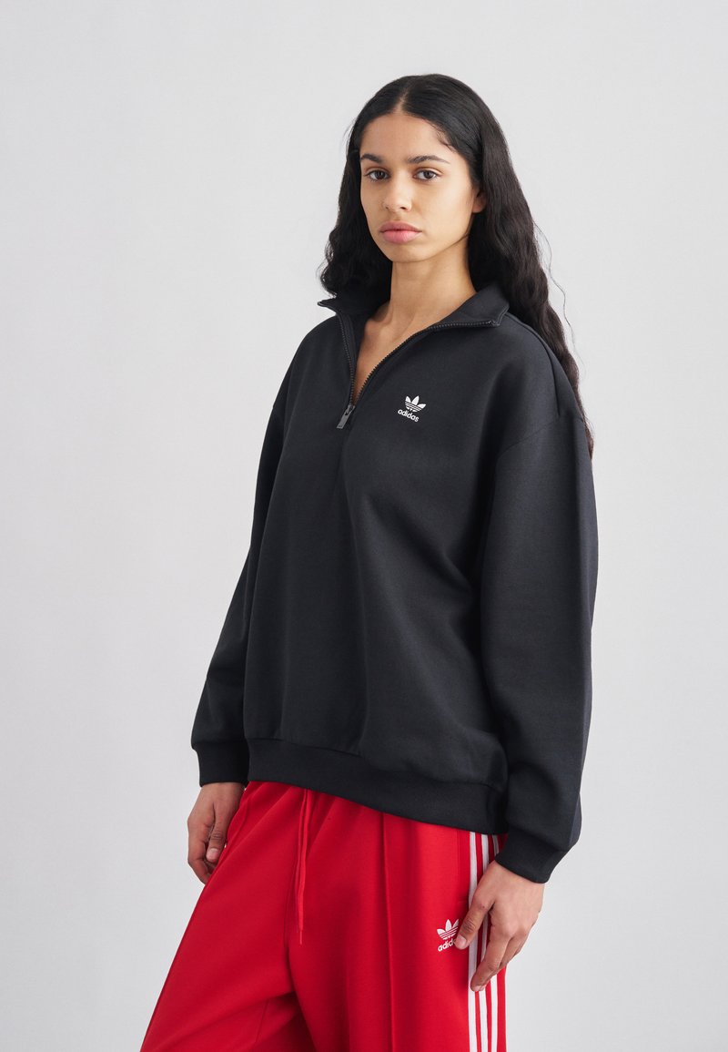 Juodas pusiau užtraukiamas vilnonis džemperis su išsiuvinėtu baltu logotipu, derinamas su raudonomis sportinėmis kelnėmis su baltomis šoninėmis juostelėmis ir atitinkančiu logotipu.