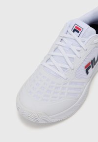 Zapatilla deportiva blanca con paneles de malla y suela de goma texturizada. Presenta un logo en rojo y azul, cordones y un sutil patrón de cuadrícula en la parte superior.