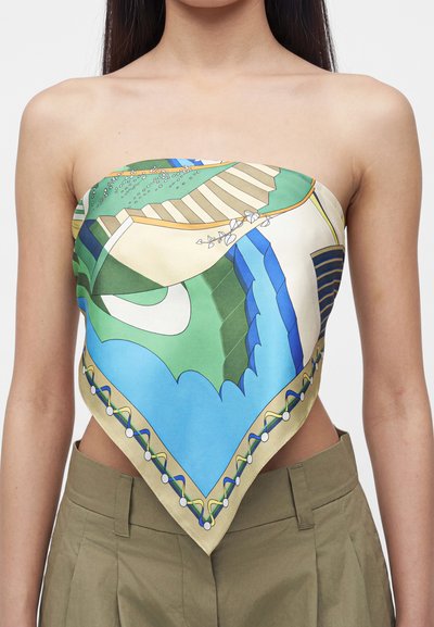 Femme portant une écharpe colorée à motifs nouée en top bustier, associée à un pantalon taille haute vert olive sur un fond uni.