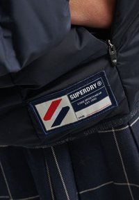 Nærbillede af en navyjakkeetiket med teksten "SUPERDRY CODE SPORTSWEAR EST. 2003" samt et rødt og blåt skrålogo og en lynlås i stof ovenfor.