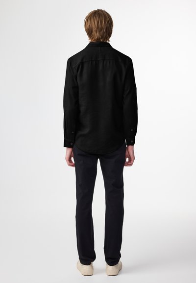 Maglietta nera a maniche lunghe con colletto, polsini con bottoni e orlo arrotondato, abbinata a pantaloni scuri e scarpe di colore chiaro.