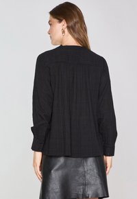 Blusa nera ampia in tessuto testurizzato, maniche lunghe, scollo rotondo e polsini con bottone, abbinata a una gonna in pelle nera.