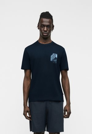 Homme aux dreadlocks courtes portant un T-shirt bleu marine avec un motif géométrique bleu clair sur la poitrine et un short bleu marine assorti.