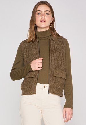 Bruine gebreide vest met een kraagontwerp, met twee voorzakken. Draagt over een donkergroene long-sleeve top en crème broek.