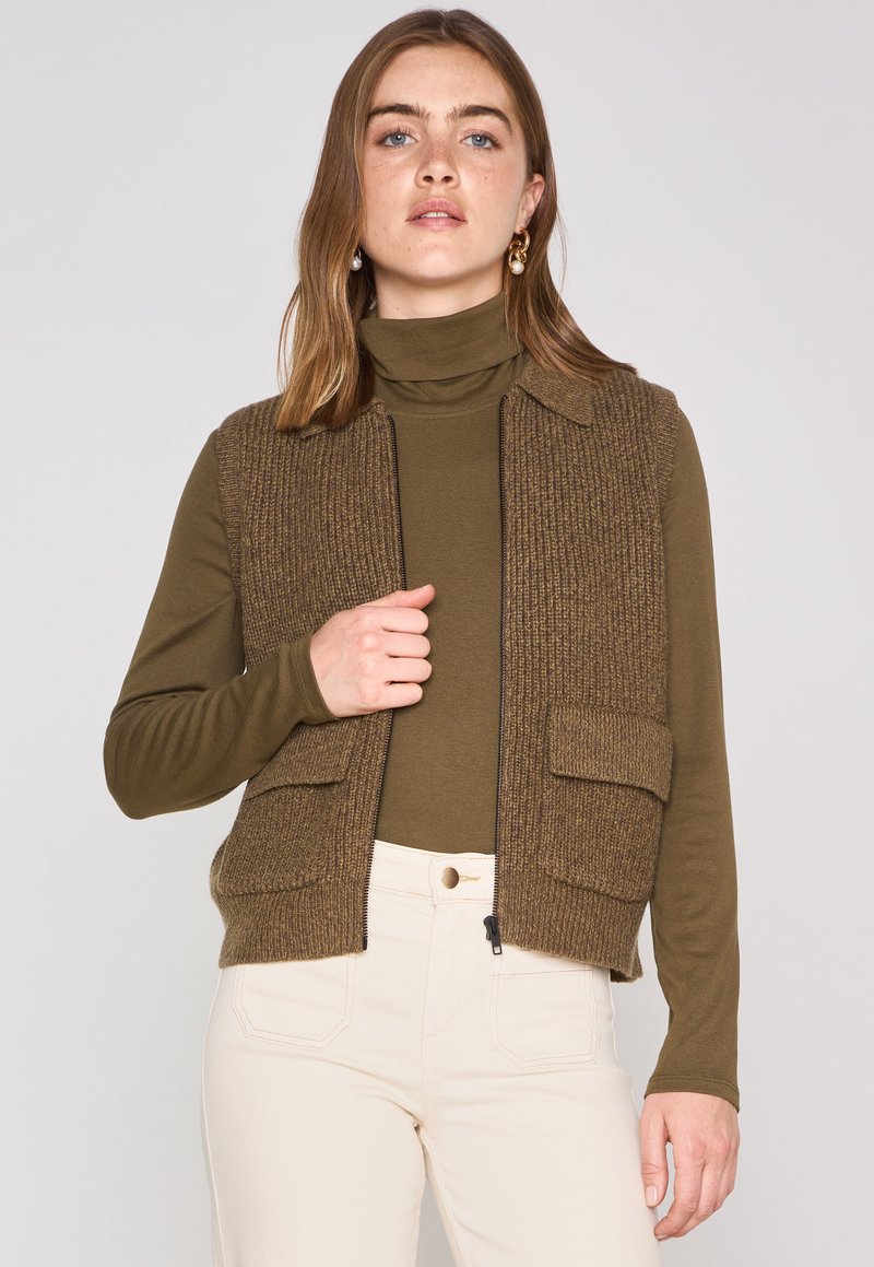 Gilet lavorato a maglia marrone con un design a colletto, dotato di due tasche frontali. Indossato sopra una maglietta a maniche lunghe verde scuro e pantaloni color crema.