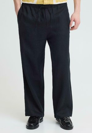 Mand iført sorte bukser med snøre og sorte loafers, stående med hænder delvist i lommer mod en ensartet baggrund.