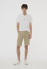 Polo bianco a maniche corte con colletto, abbinato a pantaloni corti color khaki con risvolti. Il modello indossa sneakers bianche.