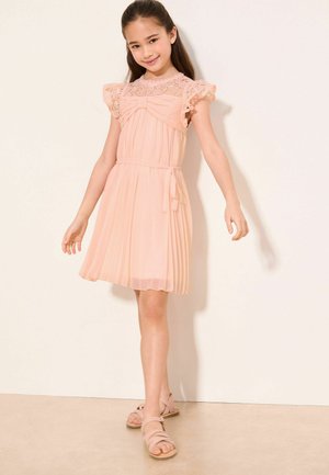 Ung pige smilende, iført en ferskenfarvet kjole med blonder og plisseringer samt en sløjfedetalje, stående foran en ensfarvet lys baggrund.