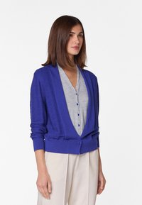 Blauwe cardigan met een diepe V-hals, lange mouwen, een textuurstof en een enkele knoop aan de voorkant. Gemaakt over een grijze top en crèmekleurige broek.
