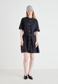 Dark linen shirt dress with button front, short sleeves, a tie waist, and a flared hem. Paired with a gray striped cap and black pointed shoes.