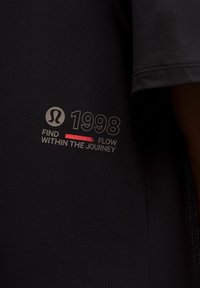 Cămașă sportivă închisă, dintr-un material texturat. Prezintă un logo, anul "1998" și textul "Caută fluxul în călătorie" cu accente gri și roșu.