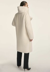 Biały wełniany płaszcz z wysokim kołnierzem, długimi rękawami i luźnym krojem, sparowany z czarnymi, dopasowanymi spodniami oraz czarnymi szpilkami w szpic.