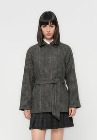 Black and white houndstooth coat with a belt, featuring a collar and long, wide sleeves. Paired with a pleated black skirt.