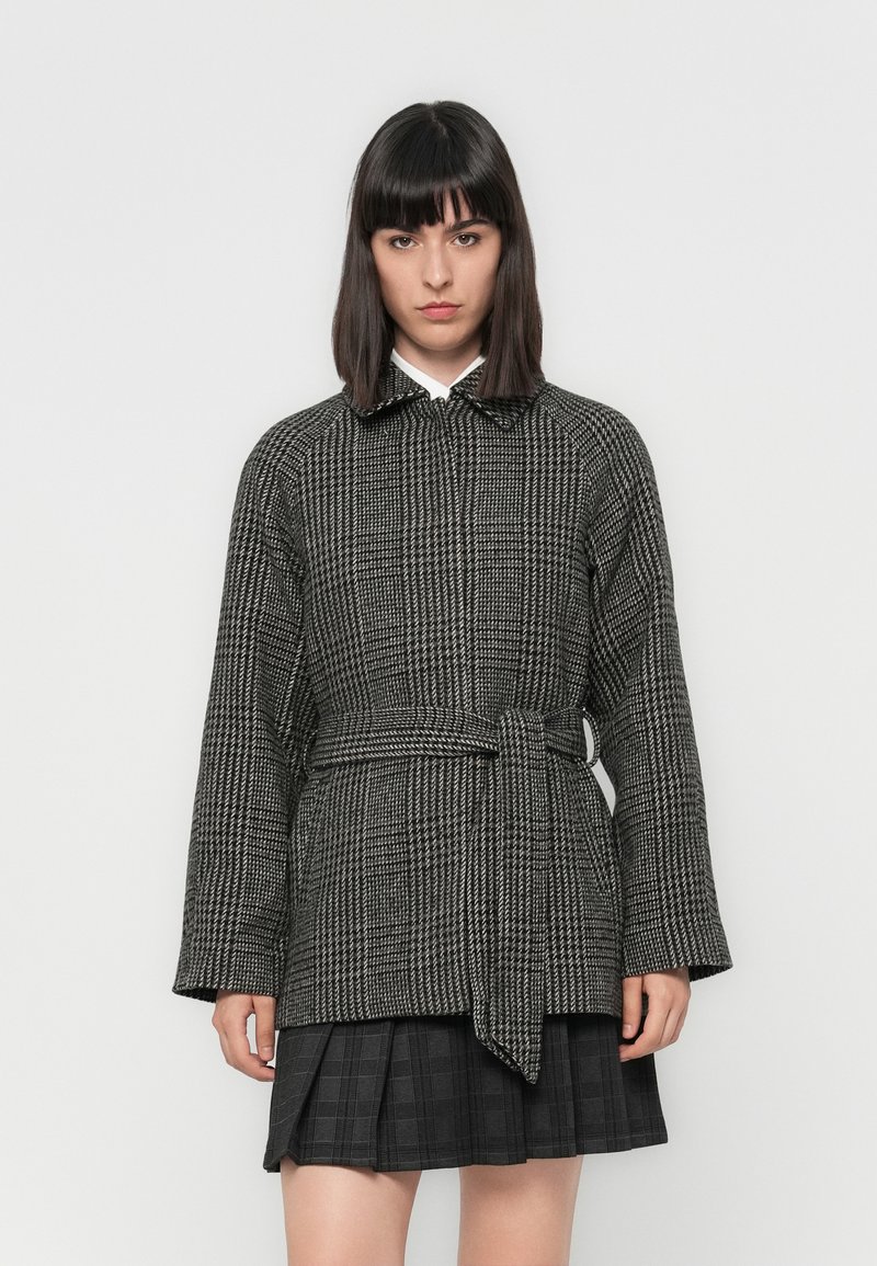 Black and white houndstooth coat with a belt, featuring a collar and long, wide sleeves. Paired with a pleated black skirt.