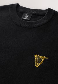 Fekete környakú pulóver apró sárga hímzett Guinness hárfa logóval a mellkasán és fekete Guinness márkajelzéssel a gallérnál.