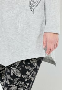 Hellgraues Oberteil mit subtiler Blattstickerei, kombiniert mit schwarzen Leggings, die weiße Blattemuster aufweisen. Fokus auf dem Saum und den Handdetails.