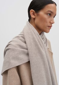 Scarf di lana grigio chiaro drappeggiato ordinatamente sulle spalle, texture morbida e finitura liscia, abbinato a un cappotto beige, caratterizzato da un design minimale.