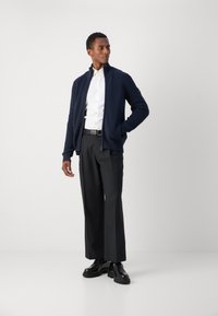 Giacca a maglia blu marina con colletto alto, abbinata a una camicia bianca e pantaloni scuri su misura. Scarpe nere con suola spessa.