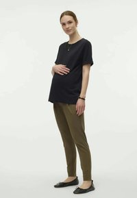 Top preto de manga curta e calças verde-azeitona com um corte descontraído. A modelo está de pé com uma mão na cintura e a outra na barriga. Sapatilhas pretas.