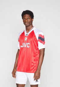 Camisa de futebol vermelha e branca com riscas verticais, apresentando o logótipo "JVC" e um emblema. Mangas curtas, acompanhada por calções brancos.