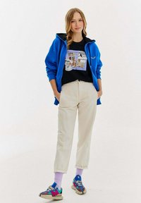 Niebieska bluzka z kapturem zapinana na zamek, na czarnej koszulce z nadrukiem, kremowe spodnie i kolorowe sneakersy z fioletowymi skarpetkami. Gładki materiał i luźny krój.