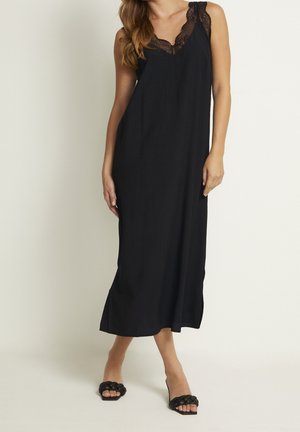 Femme portant une longue robe noire sans manches avec bordure en dentelle et des talons ouverts tissés noirs sur un fond clair.