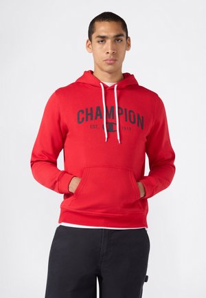 Junger Mann trägt einen roten Champion-Hoodie mit weißen Kordeln und schwarzen Hosen, steht mit den Händen in der Fronttasche vor einem einfarbigen Hintergrund.