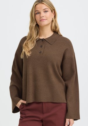 Femme aux longs cheveux blonds portant un pull ample marron à col boutonné et un pantalon bordeaux, debout devant un fond clair et uni.