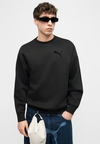 Νέος άνδρας που φοράει μαύρο φούτερ Puma και γυαλιά ηλίου, κρατώντας ένα λευκό καπέλο, με το χέρι στην τσέπη των μπλε τζιν, σε απλό φόντο.