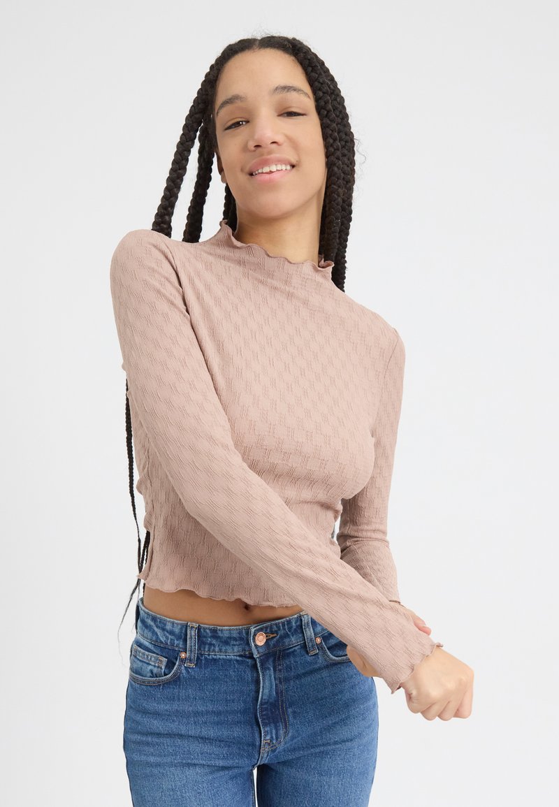 Helles rosa, strukturiertes Langarmoberteil mit Rüschensaum, croped Schnitt und gewelltem Saum. Tragen mit blauen High-Waist-Jeans.