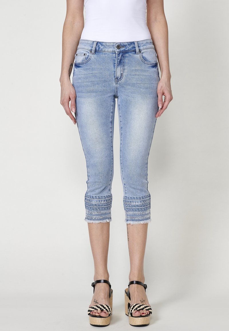 Femme portant un jean court bleu clair avec des ourlets brodés et des sandales à plateforme noires, debout devant un fond uni.