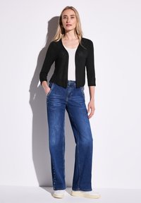 Musta neuletakki, jossa on istuva malli, yhdistettynä leveälahkeisiin sinisiin farkkuihin, joissa on metalliset yksityiskohdat, tyyliteltynä vaaleilla kengillä.