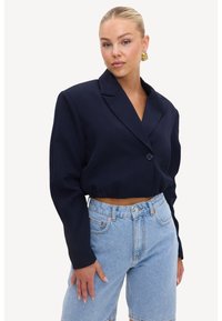 Femme aux cheveux blonds plaqués en arrière, portant des boucles d'oreilles en or, un blazer noir court et un short en jean bleu clair taille haute sur fond blanc.