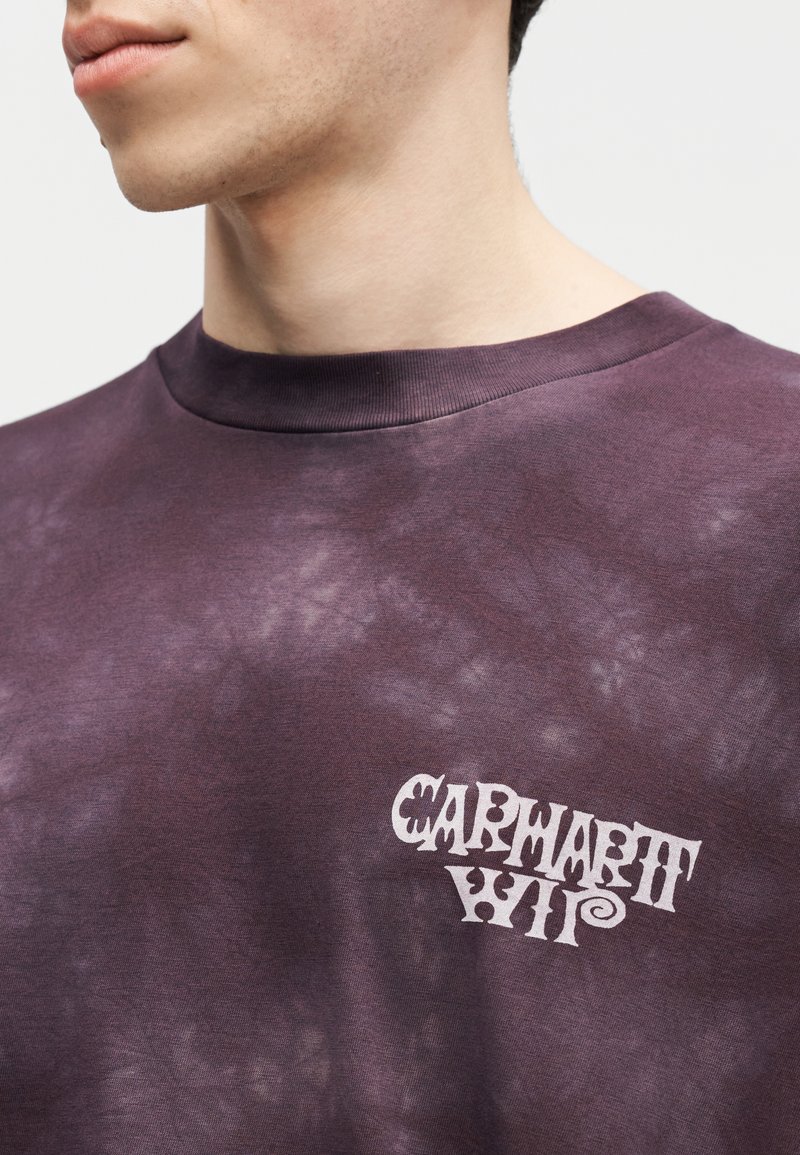 Primo piano di una persona che indossa una T-shirt Carhartt WIP tie-dye viola scuro, con il logo visibile sull'area sinistra del petto.