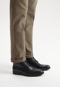Czarne skórzane botki za kostkę z okrągłym noskiem, sznurowaną konstrukcją i gładkim wykończeniem. Noszone z khaki spodniami podwiniętymi na dół.