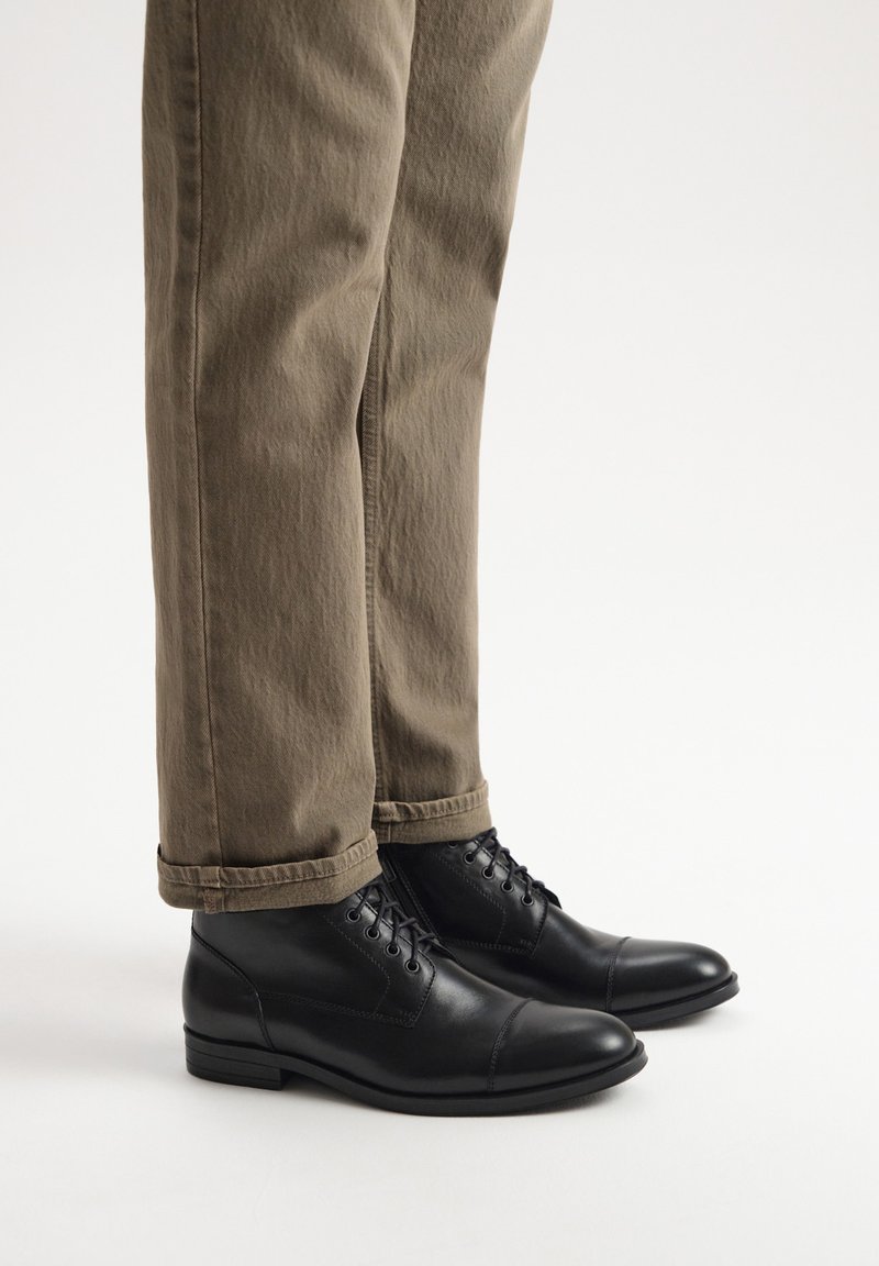 Czarne skórzane botki za kostkę z okrągłym noskiem, sznurowaną konstrukcją i gładkim wykończeniem. Noszone z khaki spodniami podwiniętymi na dół.