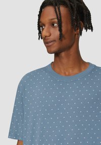 Blauwe t-shirt met korte mouwen en een ronde halslijn, voorzien van een patroon van kleine witte stippen die gelijkmatig over de stof zijn verspreid.