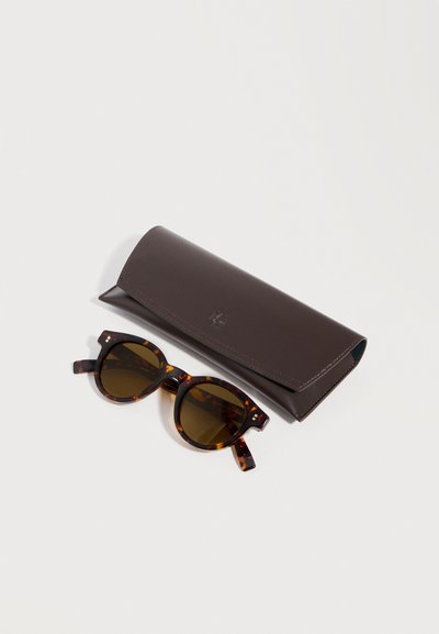Lunettes de soleil à motif écaille de tortue avec des verres ronds marron à côté d'un étui rectangulaire en cuir marron foncé.