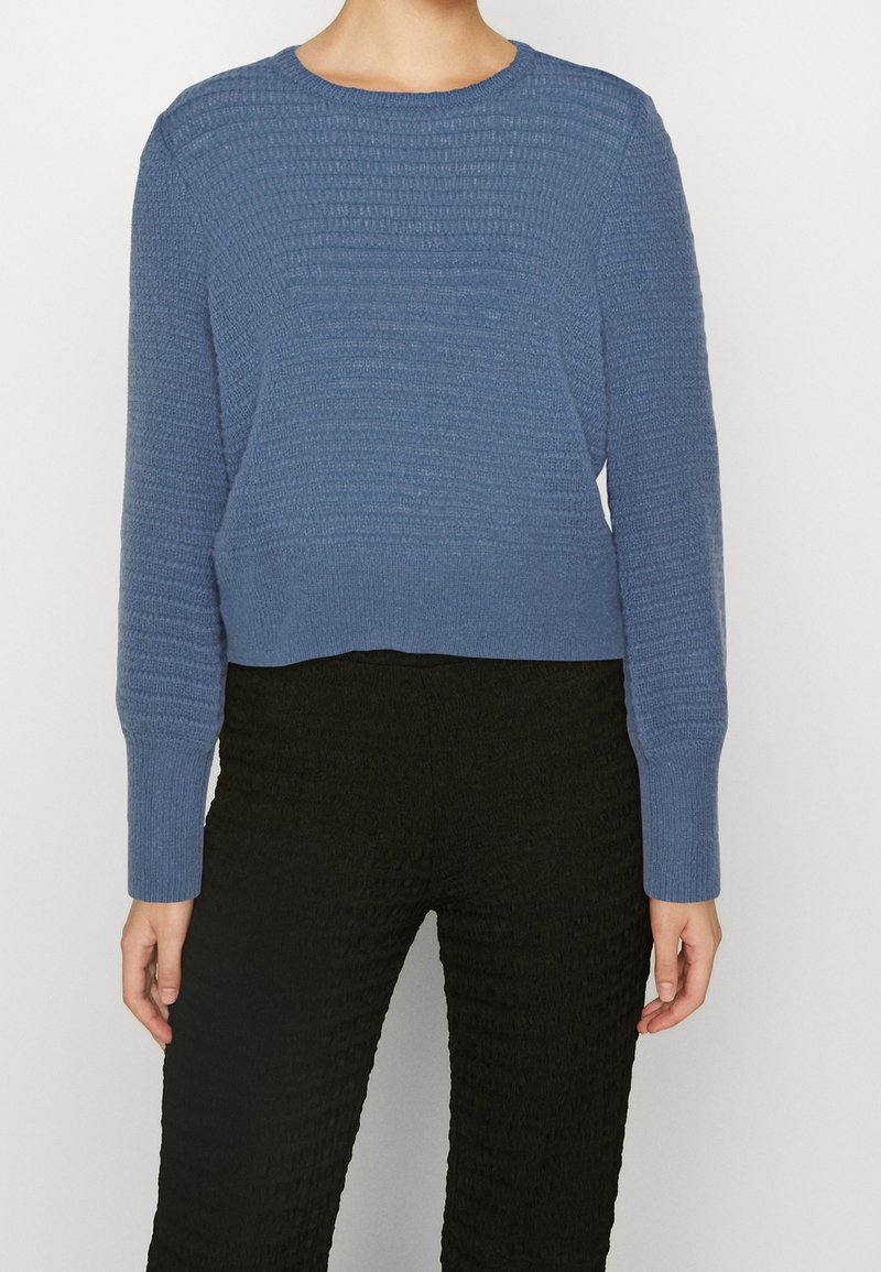 Blå texturerad tröja med rund hals och långa ärmar, med ribbat mönster och kort längd, bärs med svarta mönstrade byxor.