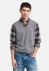 Grå ullvest över en rutig skjorta med knappar i blå, brun och vit nyanser, ihop med ljusblå jeans.