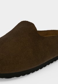Καφέ σουέντ παπούτσι slip-on με στρογγυλεμένη μύτη και μαύρη καουτσούκ σόλα, εμφανίζεται από κοντινή γωνία εμπρός-πλάι.