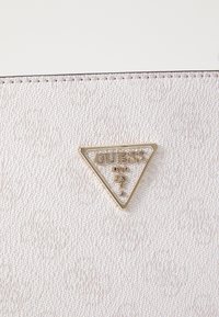Matériau texturé couleur crème arborant un logo GUESS doré en forme de triangle avec un point d'interrogation, sur fond lisse.