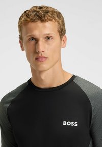Svart åtsittande topp med texturerade grå ärmar, med rund halsringning och en vit "BOSS"-logotyp på bröstet. En elegant sportig design.