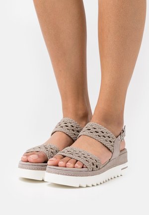 Pieds portant des sandales à brides tressées beige avec semelles compensées blanches, présentés sur un fond blanc uni.