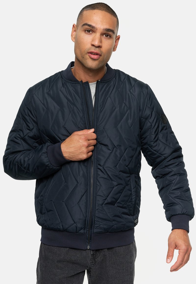 Chaqueta bomber acolchada en color azul marino con cuello redondo, cierre frontal y puños de canalé. Presenta una superficie texturizada y un detalle de logo en la manga.